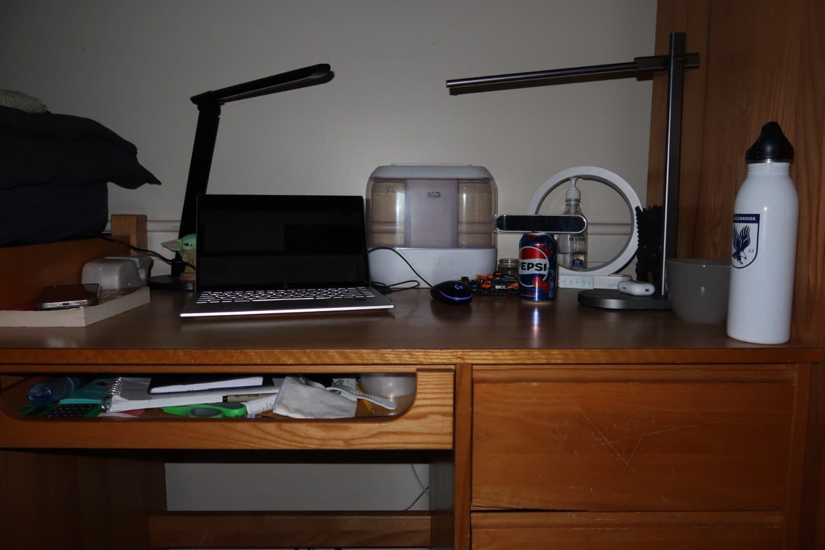 A dorm desk at night with a laptop, a lamp, and notebooks on the shelf.